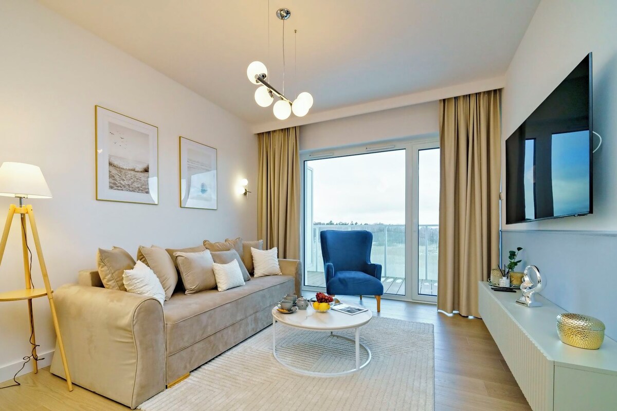 Una sala moderna con un sofá, una mesa de centro y una silla. Hay una lámpara, un televisor y una ventana con cortinas.
