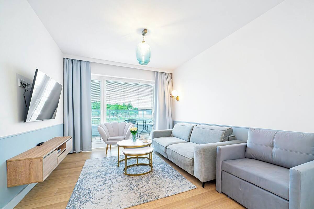 Un soggiorno moderno con un divano grigio, un tavolino rotondo e un tappeto blu decorato. Si vede una finestra con persiane, una televisione a parete e una lampada a sospensione.