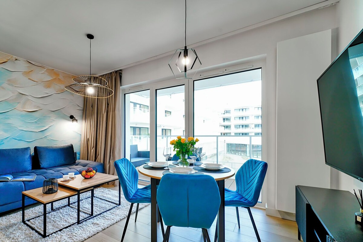 Una sala de estar moderna con un sofá azul, una mesa de comedor con sillas, un ventanal con vistas a edificios y un televisor de pantalla plana.