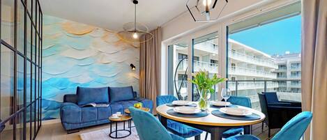 Una sala de estar moderna con un sofá azul, una mesa de comedor redonda con sillas y vistas a un edificio a través de amplios ventanales.