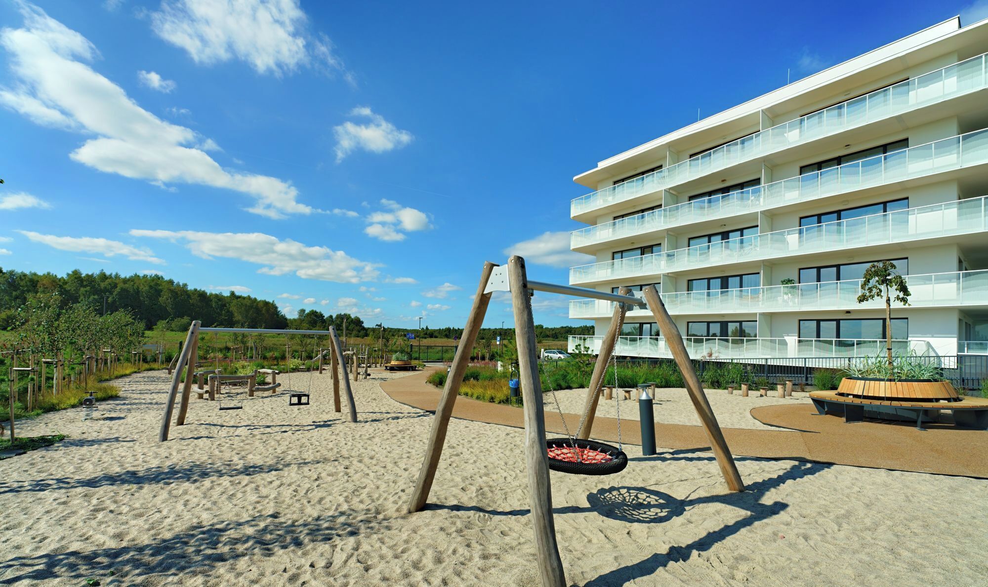 A playground with swings and a sandbox, surrounded by a building with balconies and a forested area in the background.