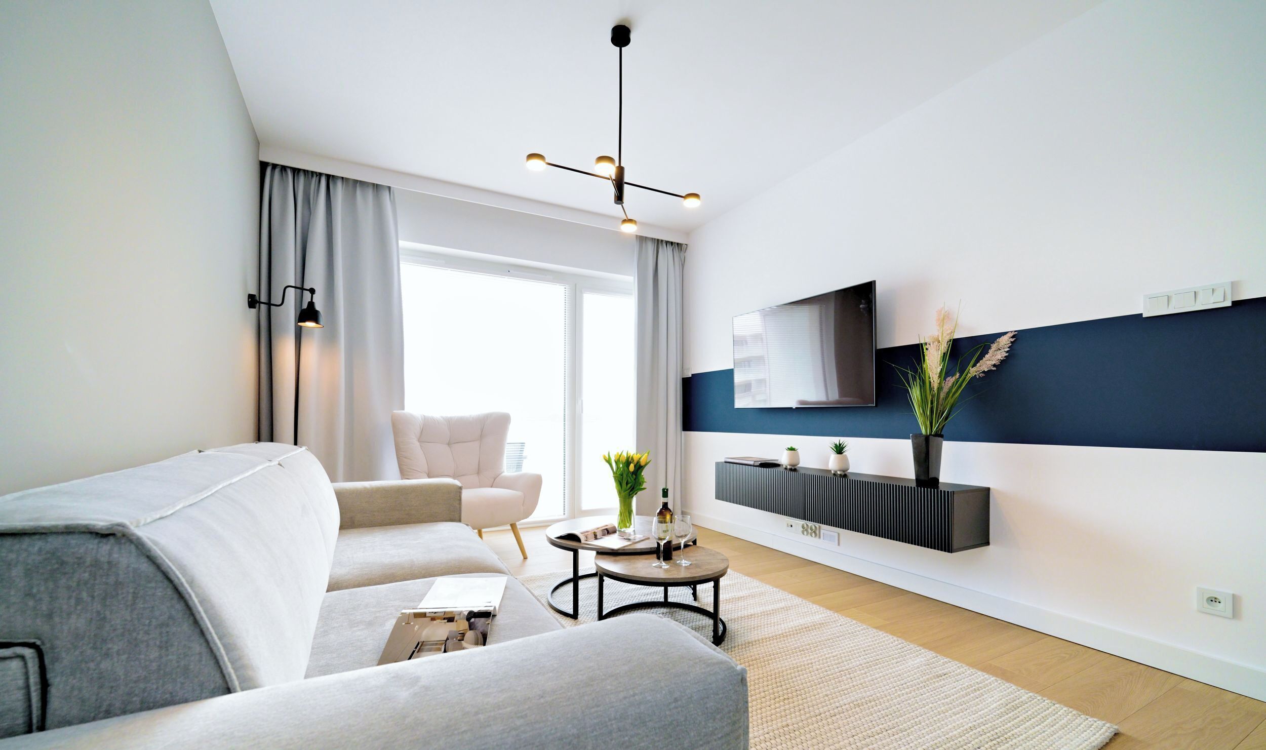 A modern living room with a sofa, a coffee table, a TV mounted on the wall, and a large window with curtains.