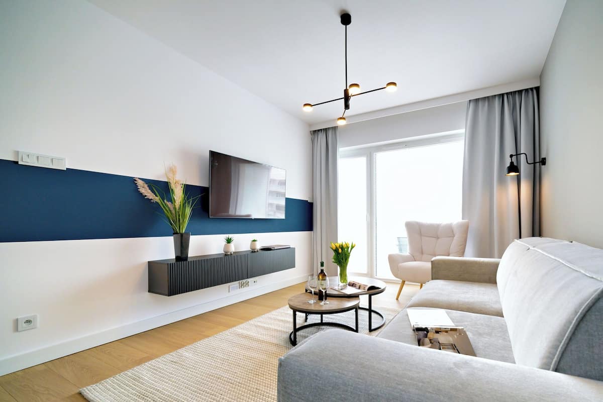 Un soggiorno moderno con un divano grigio, un tavolino, una TV a schermo piatto montata a parete e un'ampia finestra con tende.
