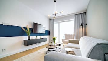 A modern living room with a grey sofa, a coffee table, a flat-screen TV mounted on the wall, and a large window with curtains.