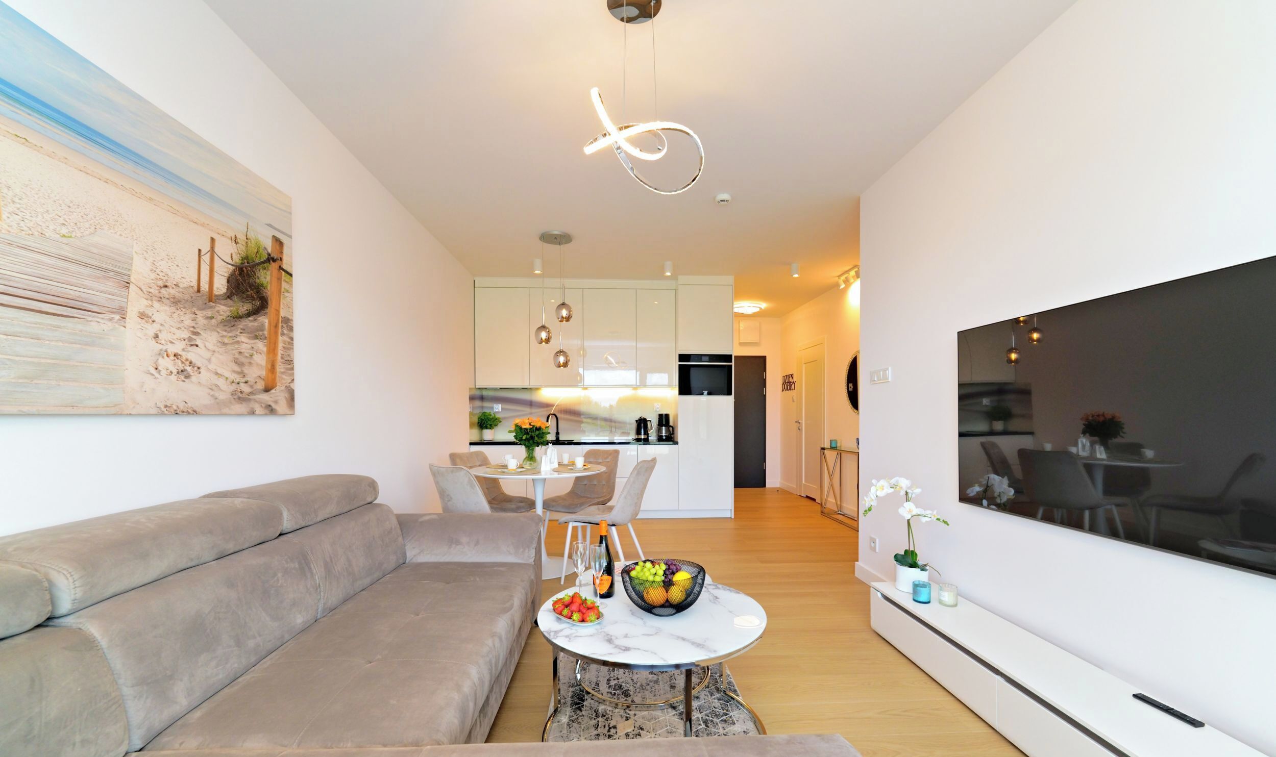 A modern living room with a grey sofa, a dining area with a white table and chairs, a kitchen in the background, and a large TV on the right wall.