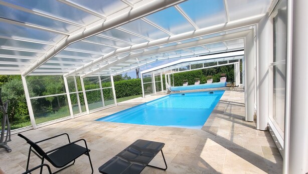 Una piscina exterior cubierta con paredes de vidrio transparente, sillas de descanso negras y suelo de baldosas.