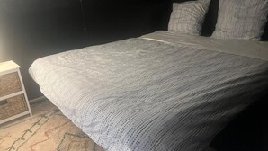 Ein Bett mit weißer Strickdecke, ein kleiner Nachttisch mit einem gewebten Korb und ein gemustertes Teppich.