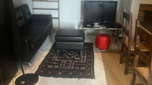 Ein Wohnzimmer mit einer schwarzen Couch, einem Fernseher auf einem Schrank, einem roten Hocker und einem gemusterten Teppich.