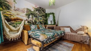 Un dormitorio con una hamaca, una cama con edredón de temática tropical, un sofá y un mural grande con una escena de bosque.