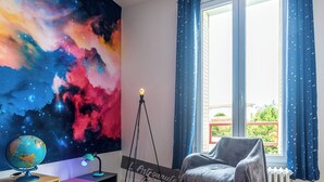 Una habitación con un mural grande con temática de galaxia, un globo en un escritorio, una silla cerca de una ventana con cortinas y una lámpara con trípode.