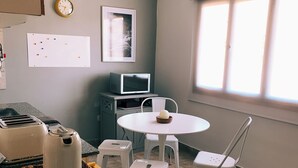 Une cuisine équipée d’un four à micro-ondes, d’un grille-pain et d’une bouilloire, le tout posé sur le plan de travail. On y trouve une table blanche avec des chaises et une fenêtre avec des stores.