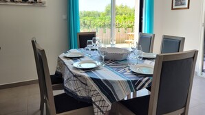 A dining area with a table set for four, chairs, a large window with curtains, and a view of greenery outside. - Spacious new house near the Casino - PLOUESCAT - beach and relaxation (Plouescat)