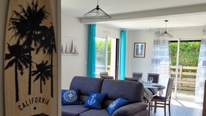 A modern living room with a grey sofa, a wooden surfboard decoration, and a dining area with a table and chairs. - Spacious new house near the Casino - PLOUESCAT - beach and relaxation (Plouescat)