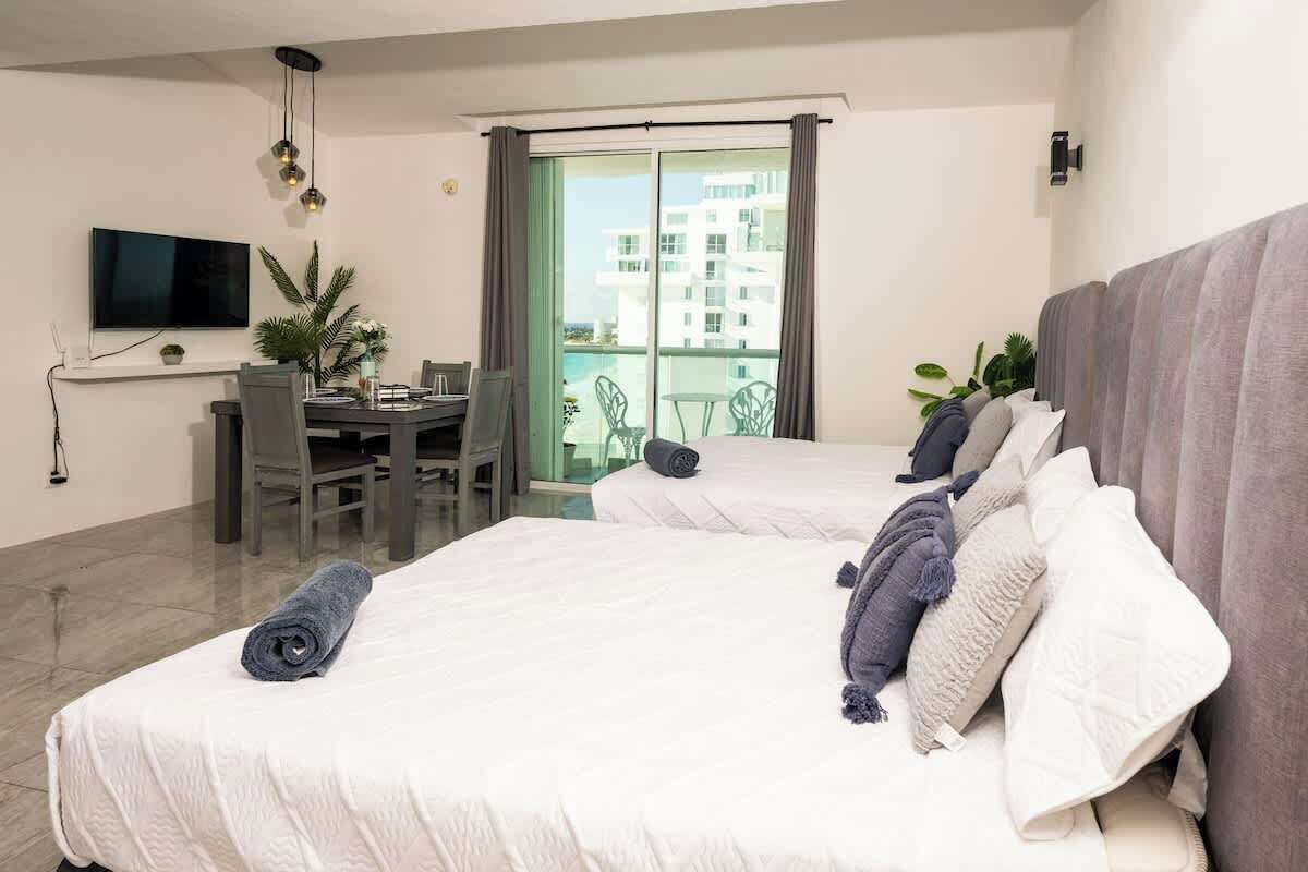 A hotel room with two beds, a dining area, a TV, and a balcony with a view of buildings.