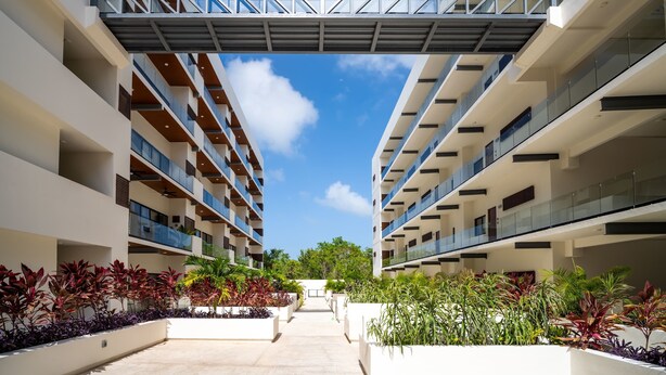 A modern multi-story building with a central walkway, surrounded by lush greenery and a clear blue sky. - Modern retreat with Rooftop Pool + Work Lounge (Cancún)