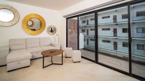 A modern living room with a sofa, ottoman, and a coffee table. There are two wall mirrors and a sliding glass door leading to a balcony. - Modern retreat with Rooftop Pool + Work Lounge (Cancún)