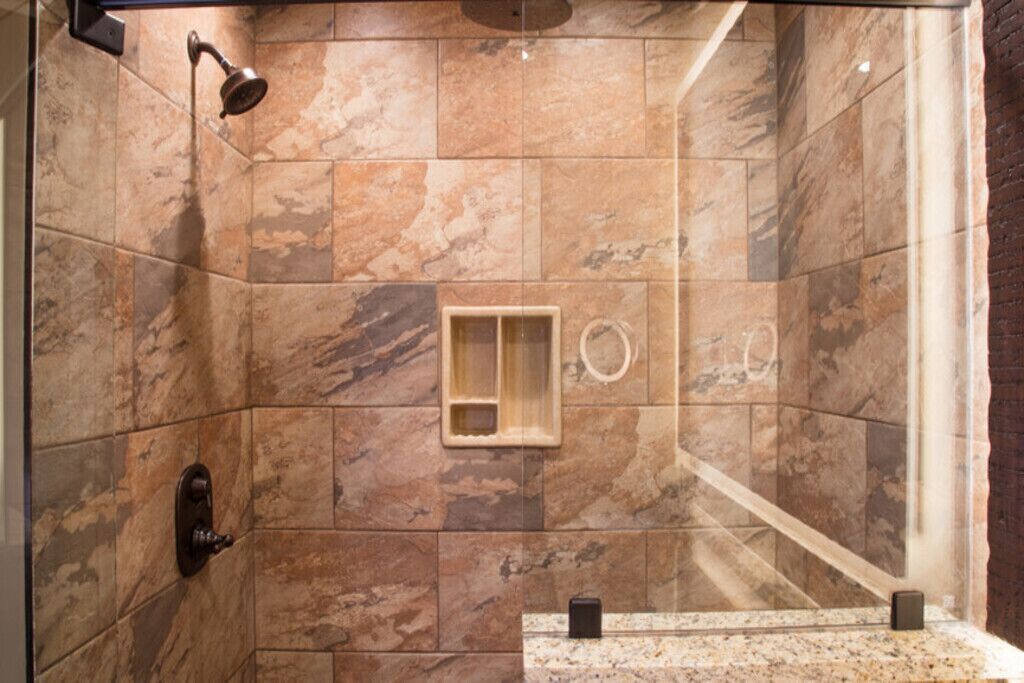 A shower area with a marble wall, a handheld showerhead, a small wooden shelf, and a glass shower enclosure.