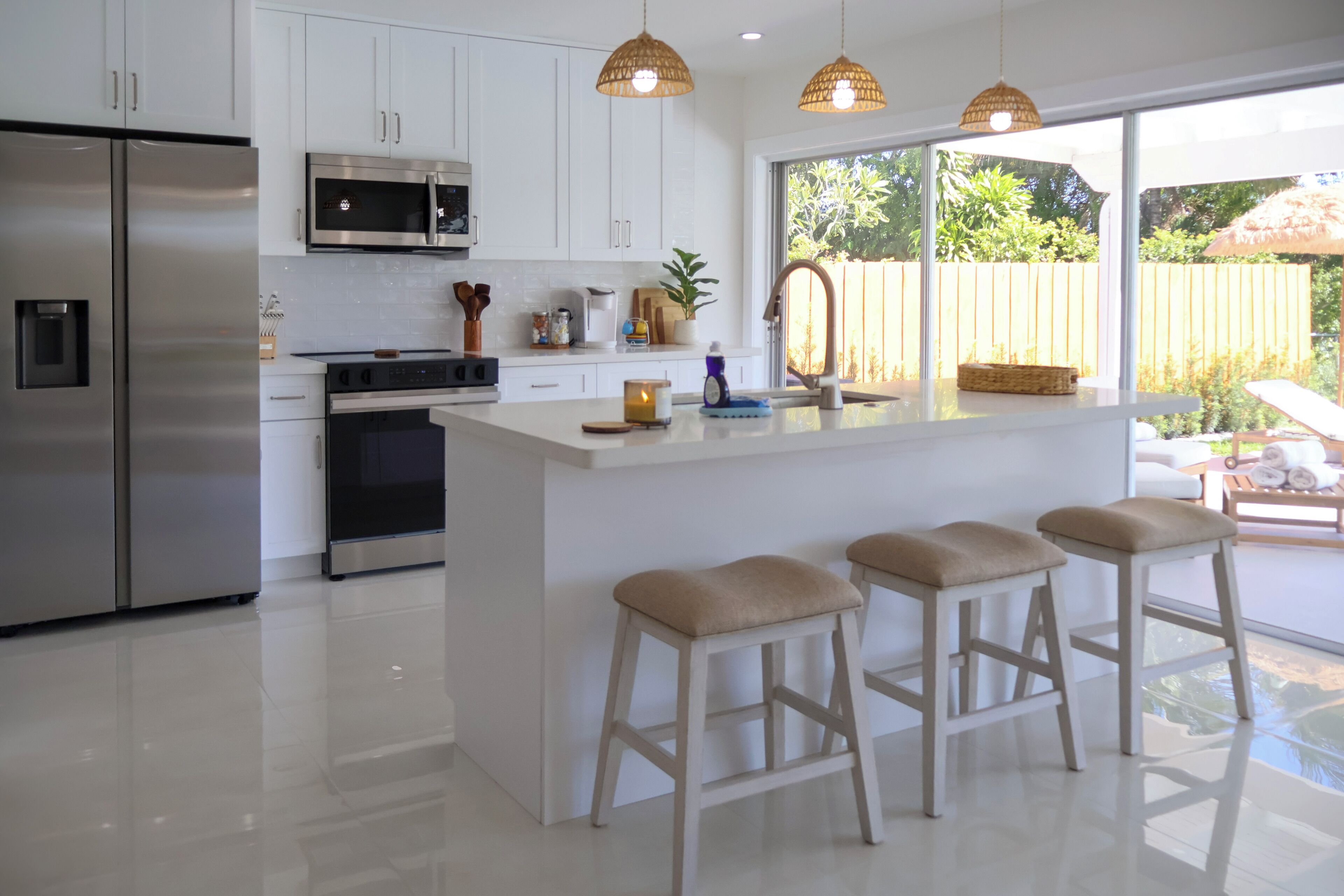 Une cuisine moderne dotée d’appareils électroménagers en acier inoxydable, d’un grand îlot central avec des tabourets, et offrant une vue sur le jardin arrière grâce à une porte-fenêtre coulissante.