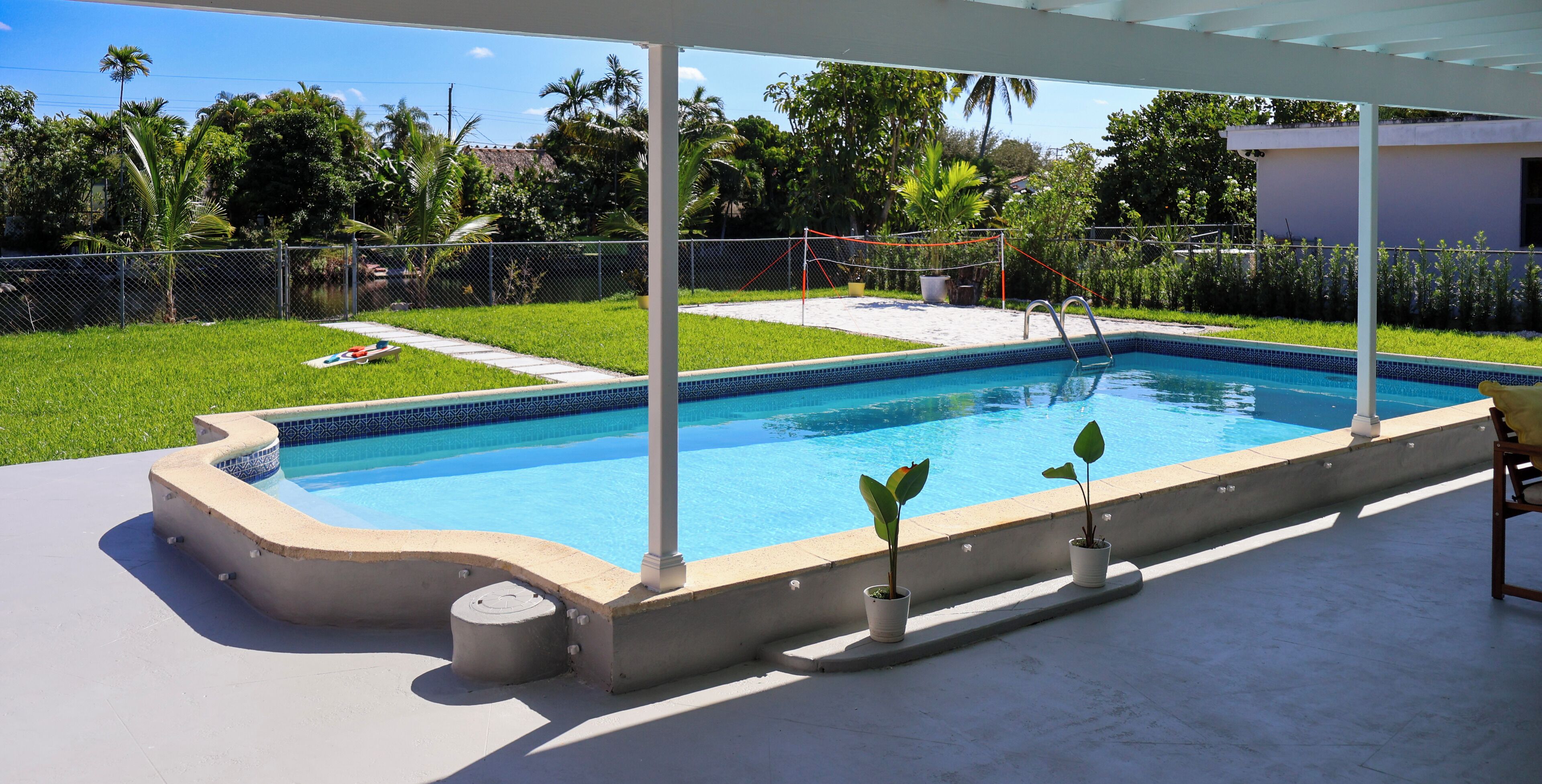 Uma piscina retangular com borda revestida, rodeada por um deck de concreto. Há duas plantas em vasos no deck e uma escada de metal para entrar na piscina.