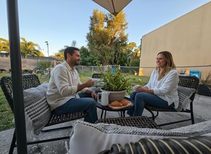 Two people seated at an outdoor table with a potted plant, a tray of pastries, and a clear view of the surrounding garden and fence. - 3BR Funhouse • Game Lounge • Near Beach & Dining in Cape Coral (Cape Coral)