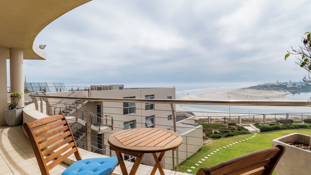 A balcony with a wooden chair and table, overlooking a beach and buildings. - Lagoon Shoreline Penthouse (Cape Town)
