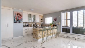 A modern kitchen with a marble floor, a central island with bar stools, and a large window providing natural light. - Lagoon Shoreline Penthouse (Cape Town)