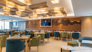 A modern restaurant with wooden tables and teal chairs, a large TV screen, and a ceiling with geometric light fixtures. - Lagoon Shoreline Penthouse (Cape Town)