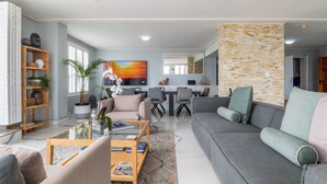 A modern living room with a grey sofa, a coffee table, and a TV. There is a dining area with chairs and a table in the background. - Lagoon Shoreline Penthouse (Cape Town)