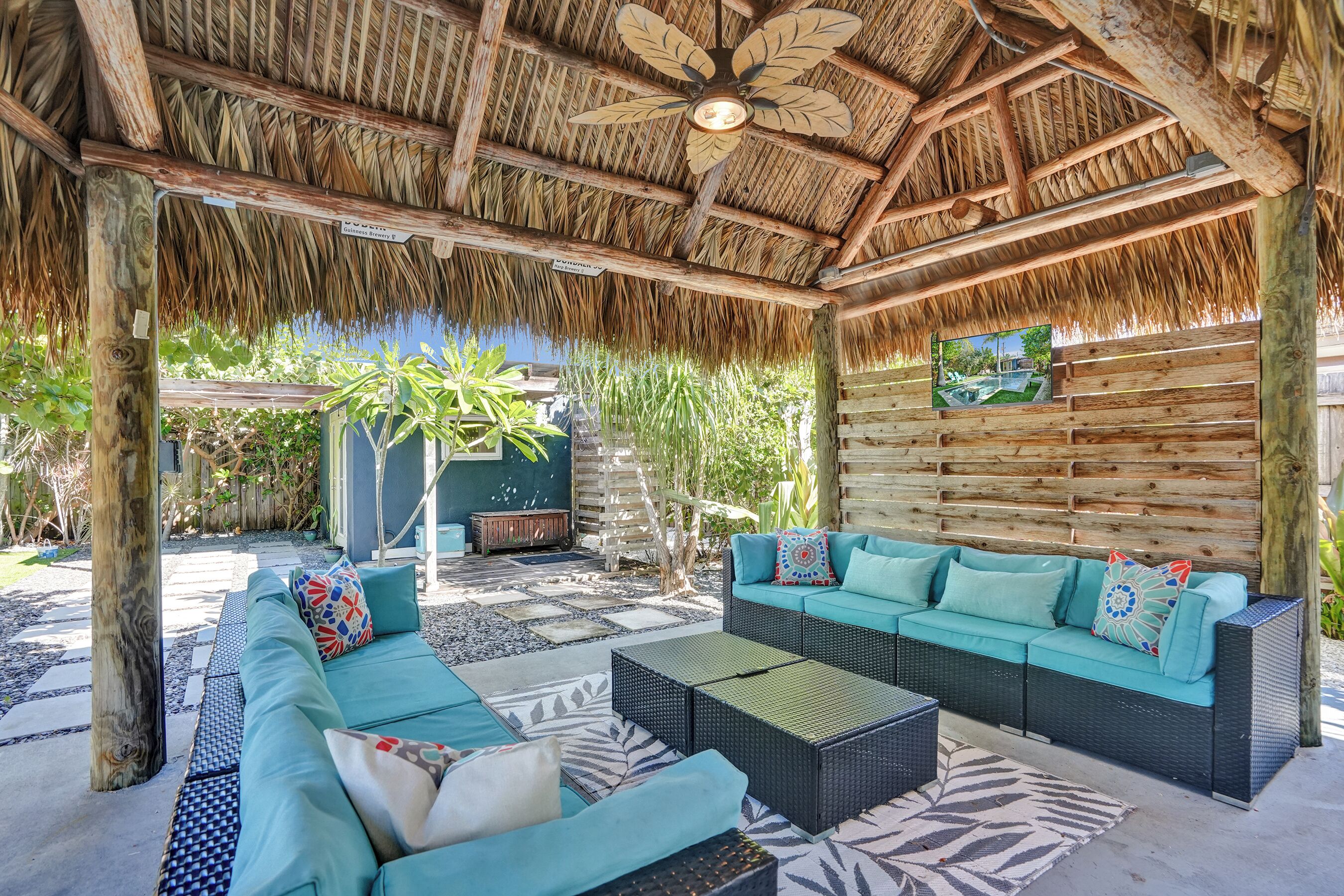 A covered outdoor seating area with a thatched roof, wicker furniture, and a wall-mounted television.