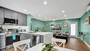 A modern kitchen with grey cabinets, a stainless steel oven, and a microwave. There is a dining area with a white table and chairs, and a living room with a sofa and a TV. - NEW Tropical Oasis with Heated Pool & Tiki Hut (Pompano Beach)