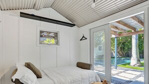 A bedroom with a bed, a small table with a decorative item, a window with a view of a garden, and a hanging light fixture. - NEW Tropical Oasis with Heated Pool & Tiki Hut (Pompano Beach)