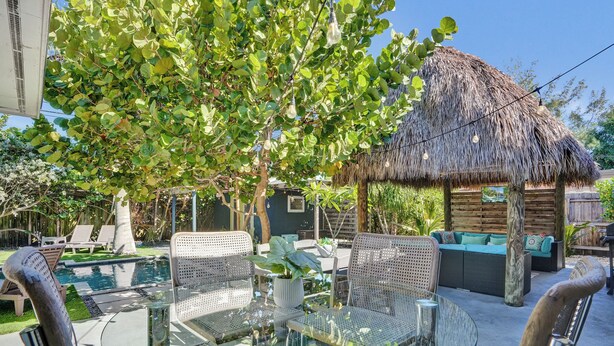 A glass-top table with wicker chairs, a thatched-roof structure, and a pool in the background. - NEW Tropical Oasis with Heated Pool & Tiki Hut (Pompano Beach)