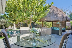 A glass-top table with wicker chairs, a thatched-roof structure, and a pool in the background.