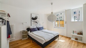 Un dormitorio con cama, un radiador, un armario, una lámpara colgante y una ventana con vista a edificios.