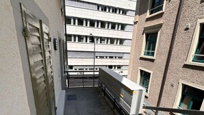 Un corridoio stretto con pavimento in cemento, ringhiera in metallo e vista su un edificio a più piani con finestre e un balcone.