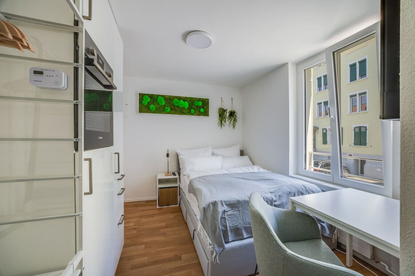 Une chambre moderne avec un lit, une table de chevet et une fenêtre donnant sur des immeubles.