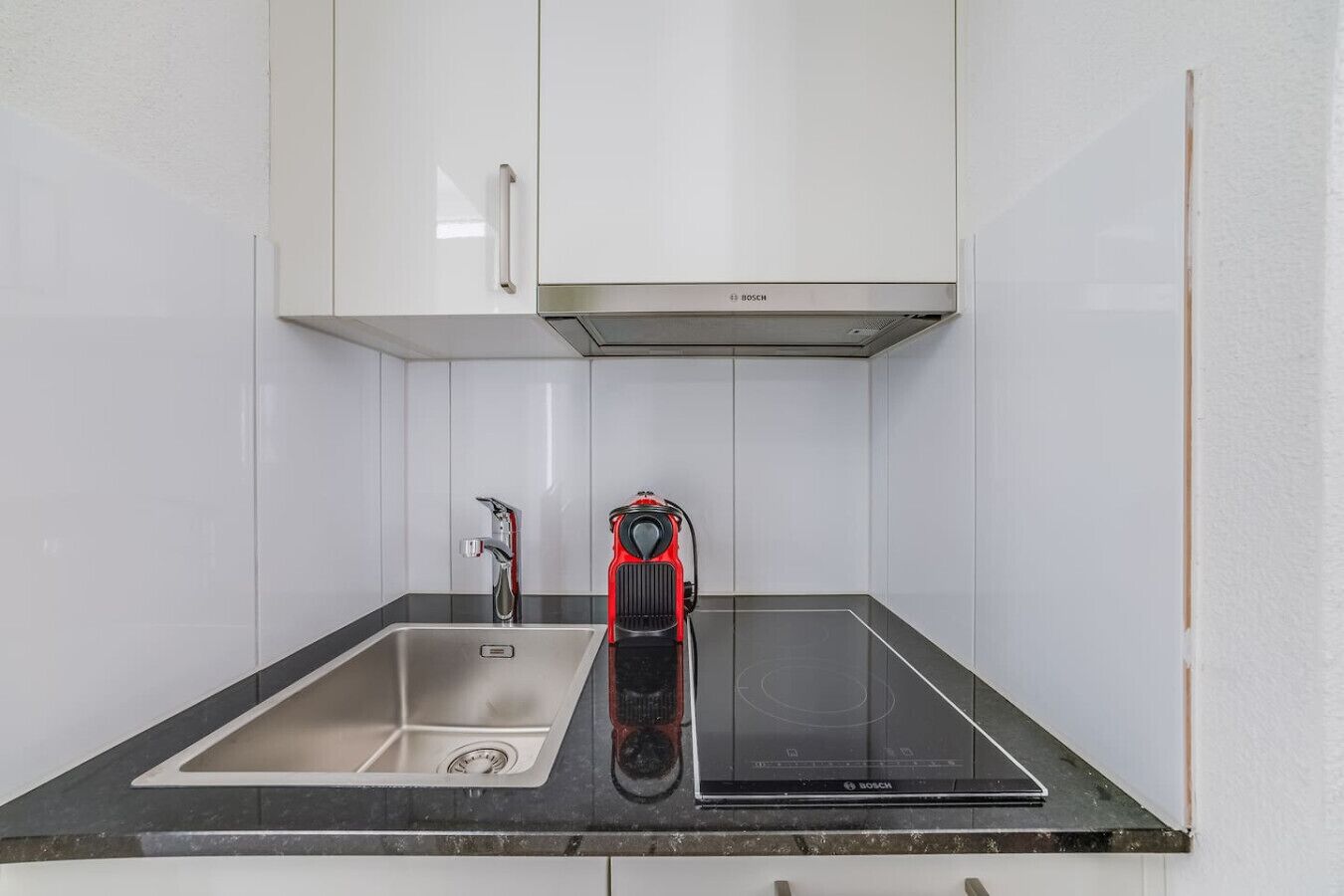 Cocina con armarios blancos, un lavamanos de acero inoxidable, una placa de inducción Bosch y un hervidor eléctrico rojo.