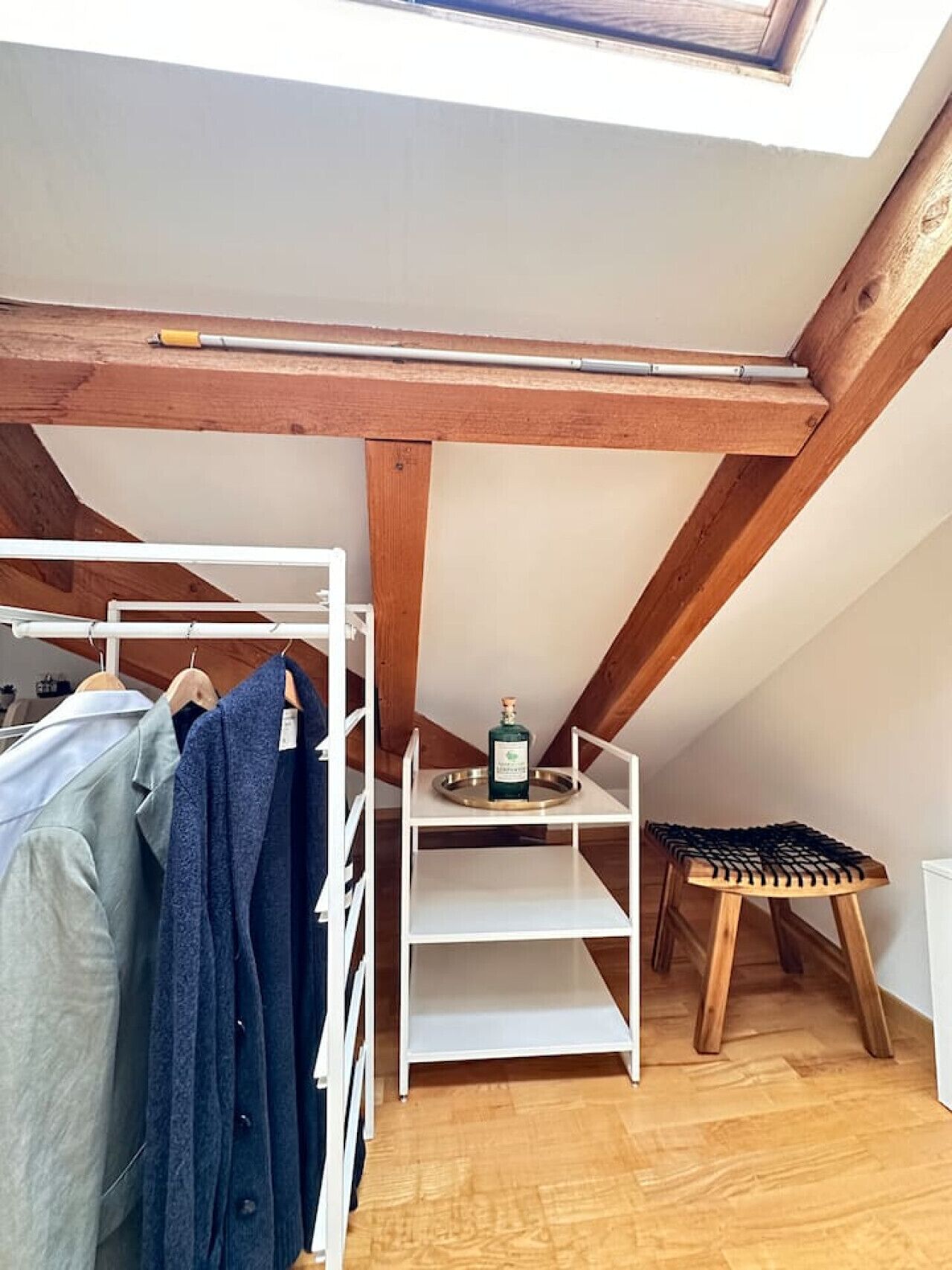 Ein Schlafzimmer im Loft-Stil mit Dachfenster, Holzbalken, einer weißen Regalkonstruktion, einem kleinen Hocker und einer Kleiderstange.