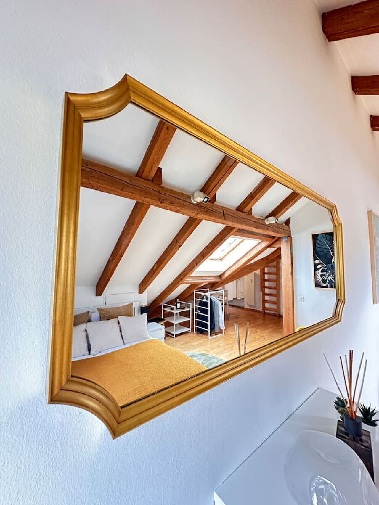 A room with a large mirror, wooden beams, a bed, a white dresser, and a small plant.