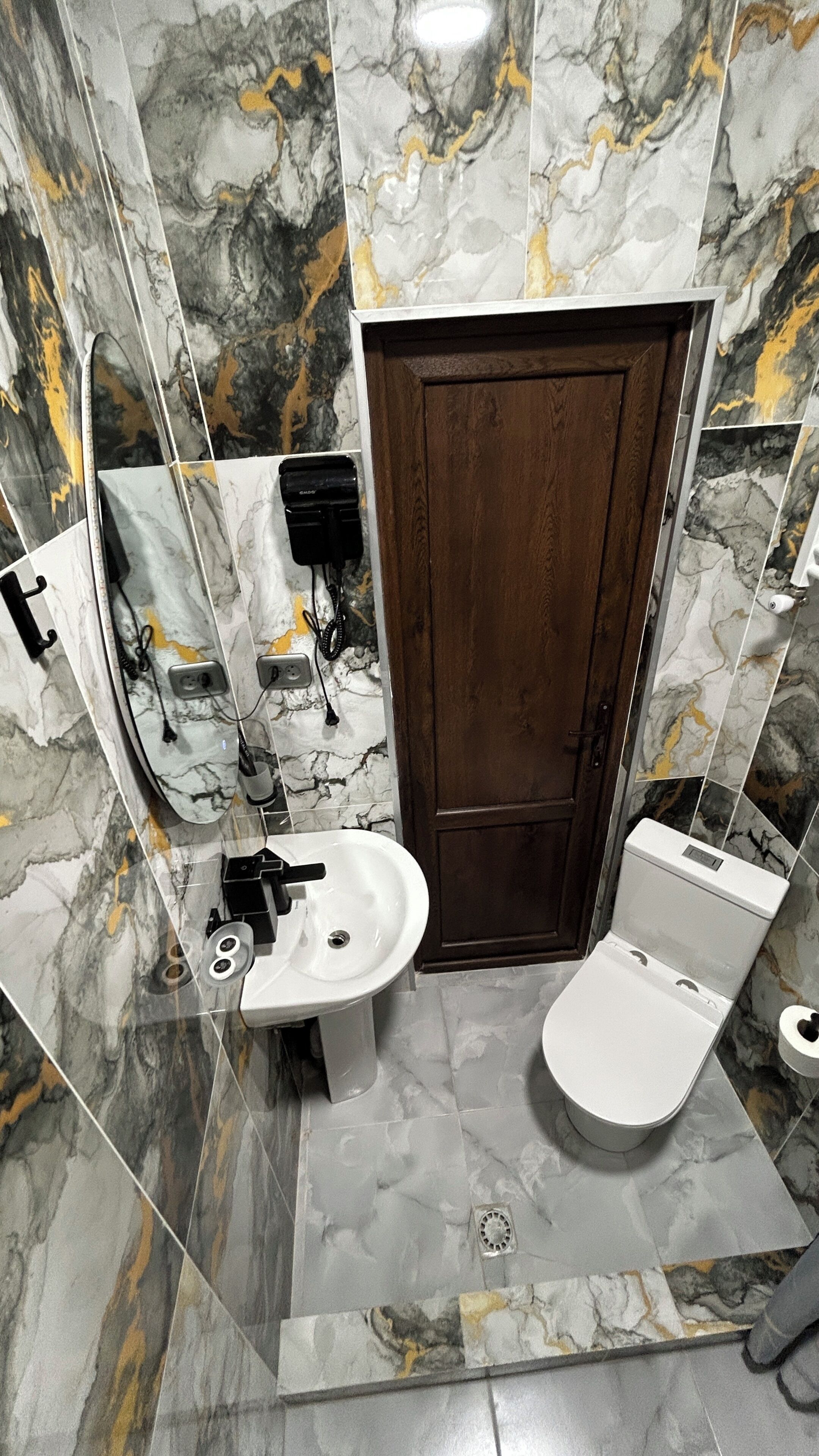 Een badkamer met een marmeren muur, een houten deur, een witte wastafel en een toilet.
