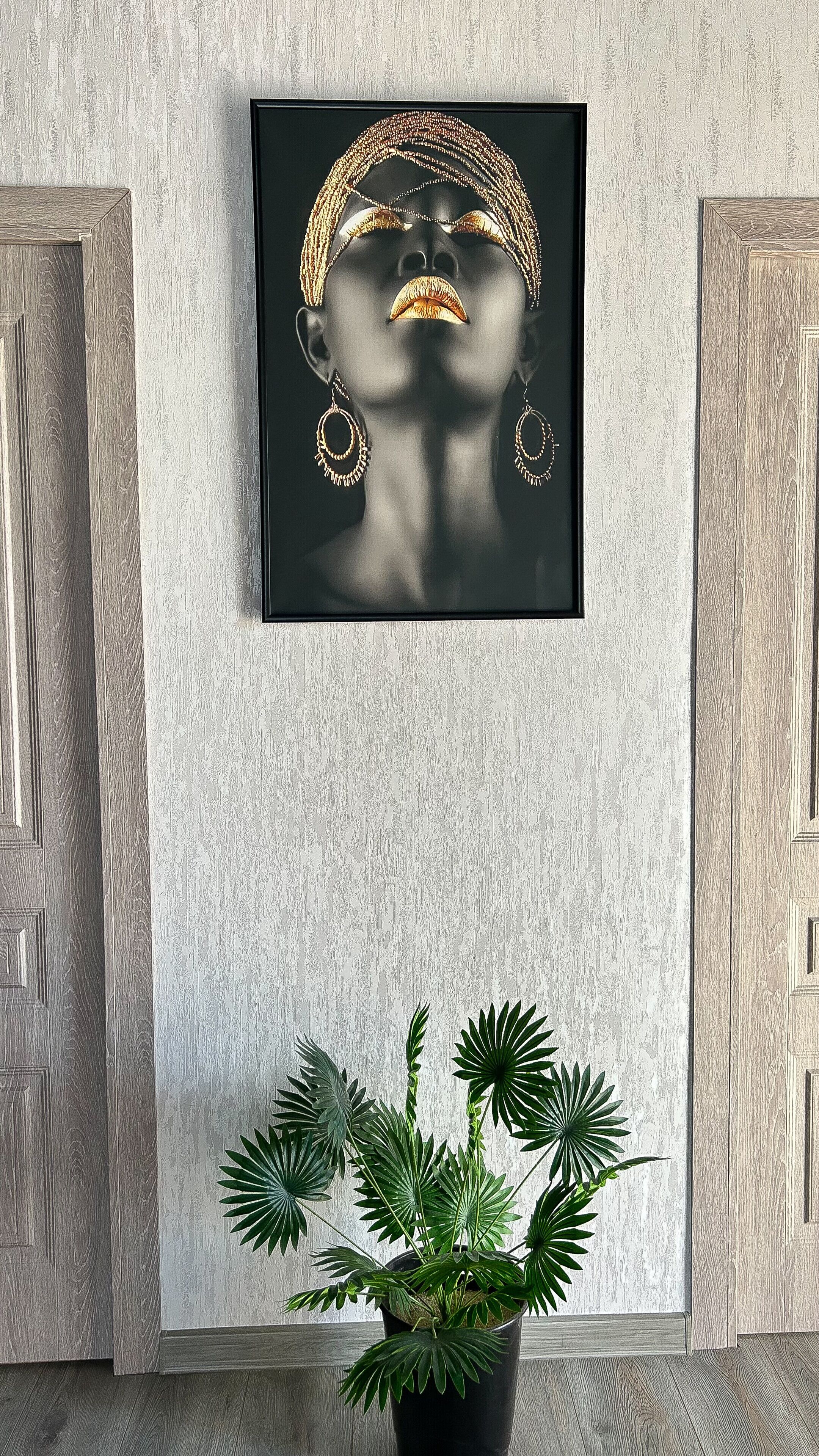 Una obra de arte enmarcada que muestra a una mujer con joyas en una pared, una planta en maceta debajo y puertas de madera a cada lado.