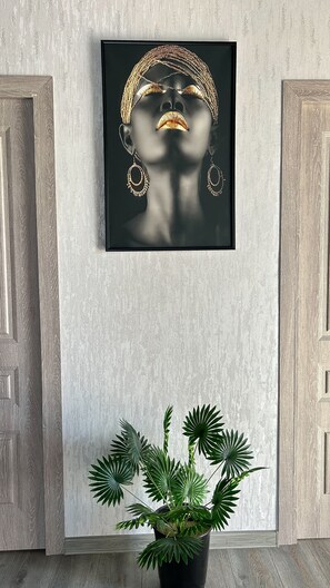 Un'opera d'arte incorniciata raffigura una donna con gioielli, appesa a una parete, con una pianta in vaso sottostante e porte in legno ai lati.