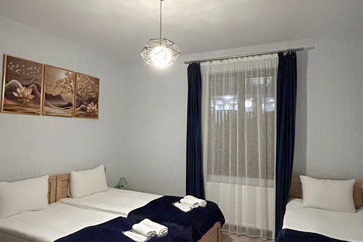 Ein Hotelzimmer mit zwei Einzelbetten, Holzboden, einer Deckenleuchte und einem Fenster mit durchsichtigen Vorhängen.