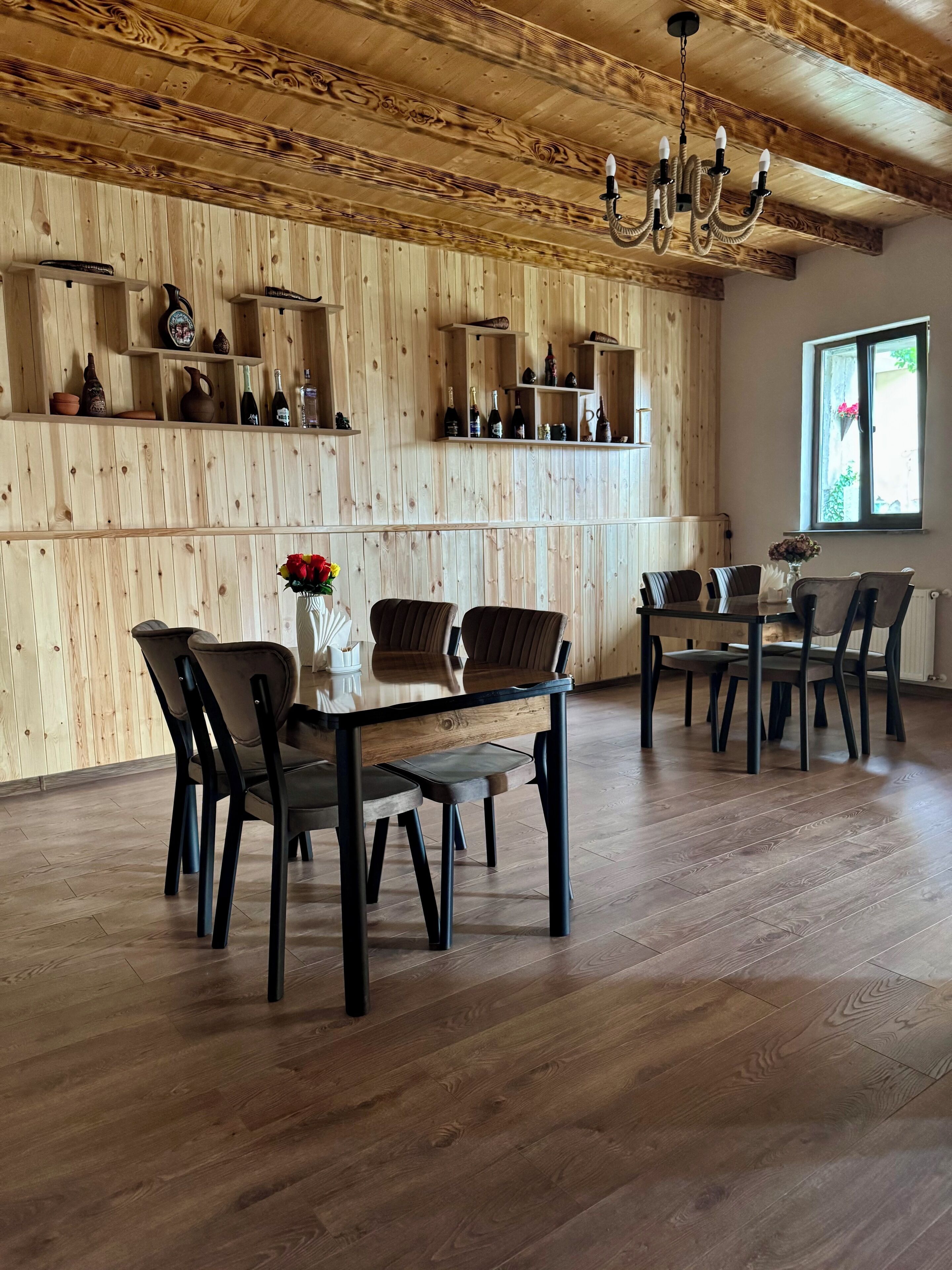 A wooden-paneled room with a dining table and chairs, a chandelier, and shelves with bottles and pottery.