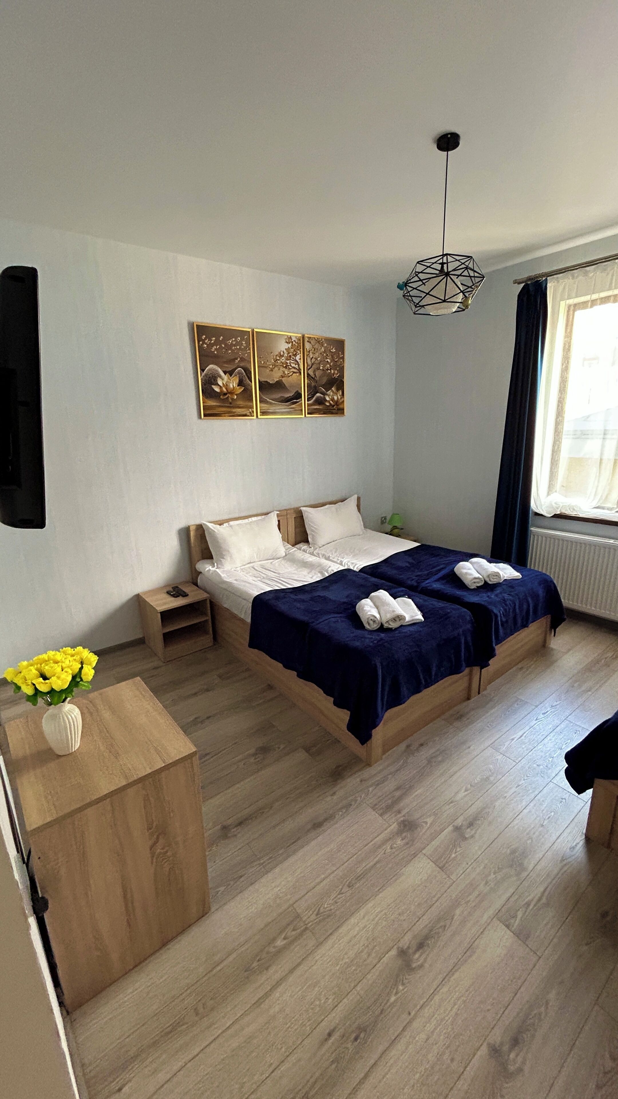 A bedroom with a bed, bedside tables, a nightstand, a vase with flowers, and a window with curtains.