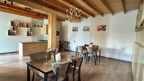Een ruime kamer met houten balken, een eethoek met stoelen en een tafel, en een keuken met planken en flessen.