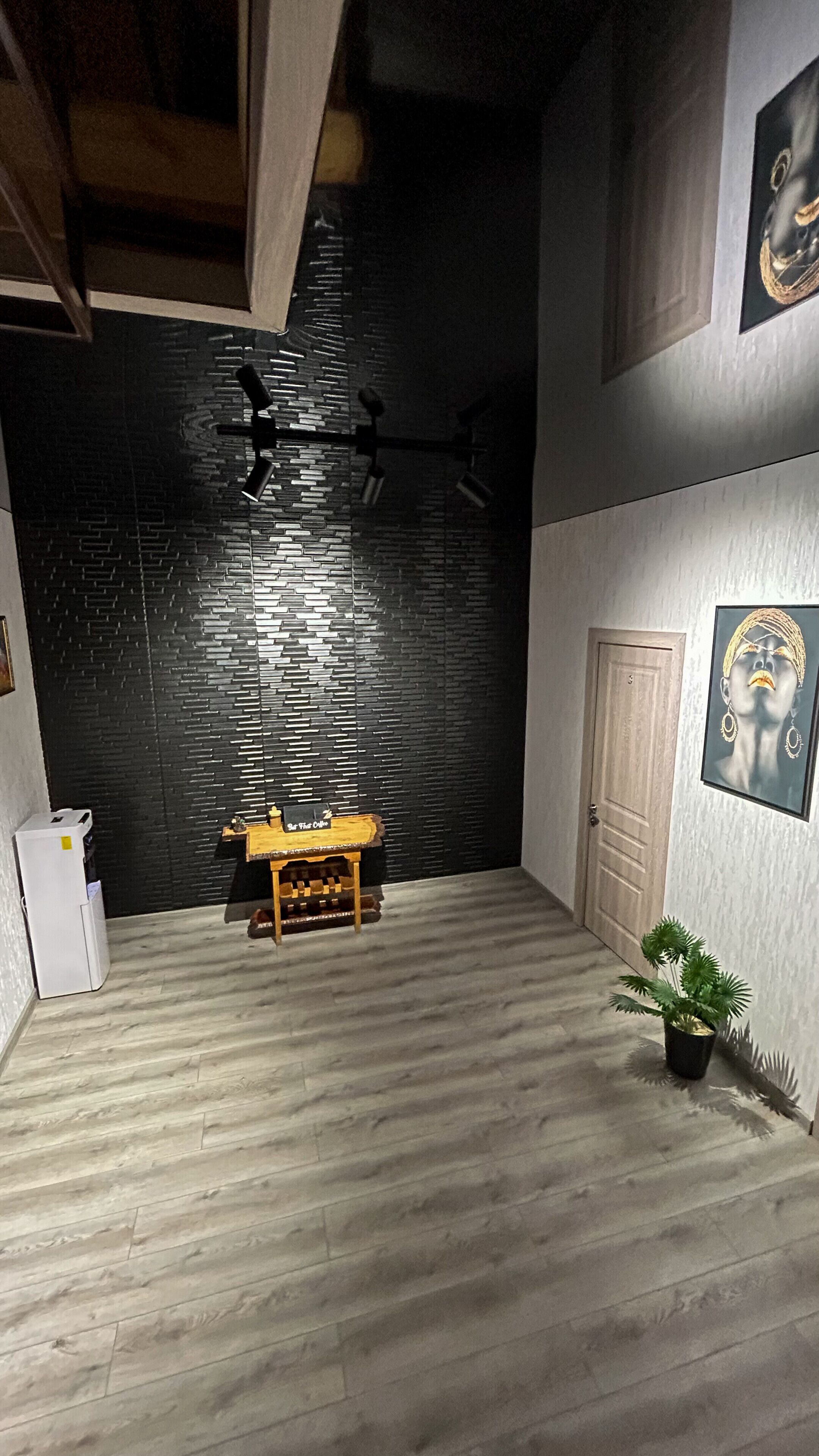 Une pièce avec un plancher en bois, un mur noir à texture, une porte en bois, une petite table, une plante en pot et un ventilateur de plafond.