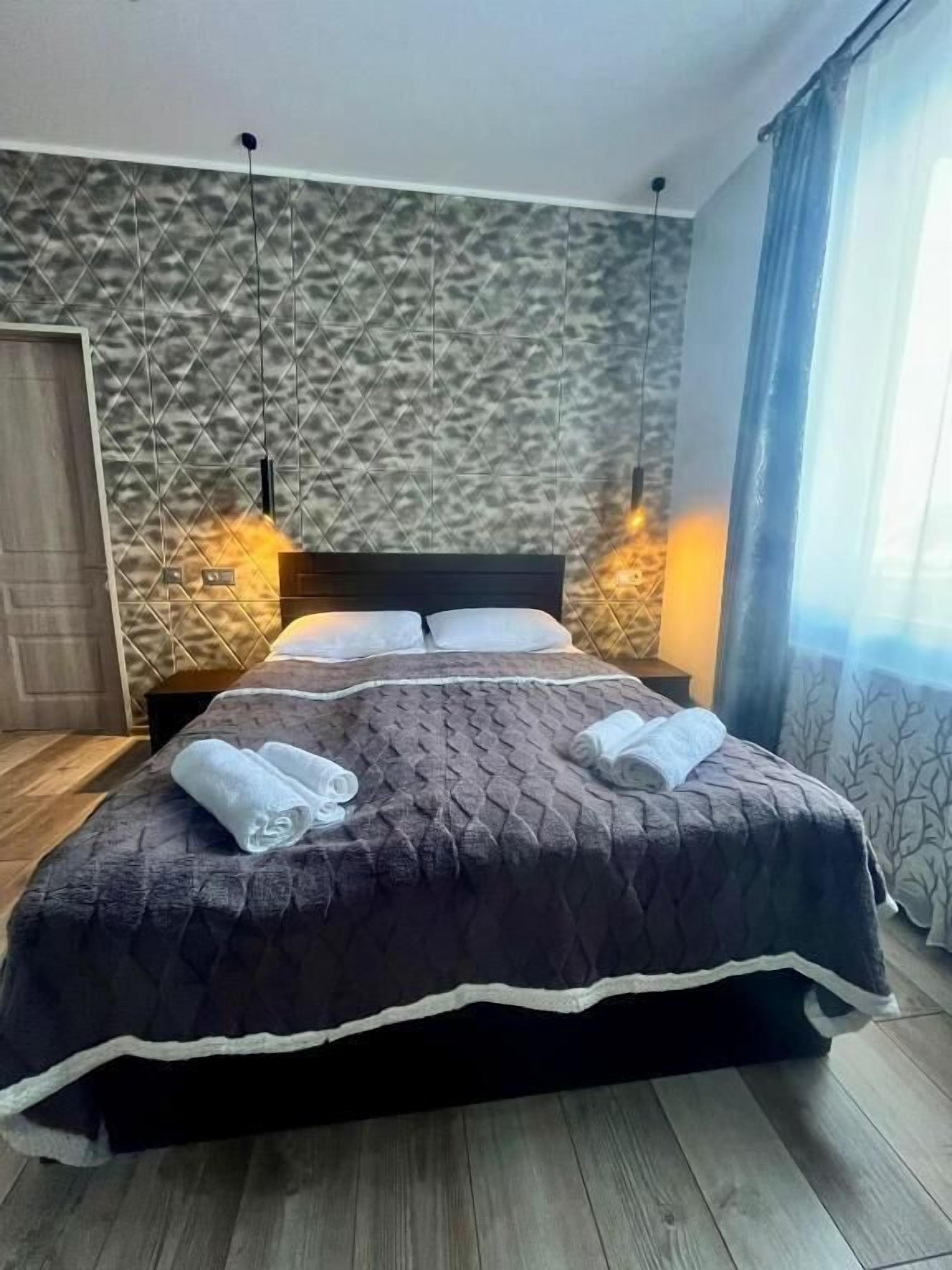A neatly made bed with a dark quilt and white trim, a textured wall behind, and a wooden floor.