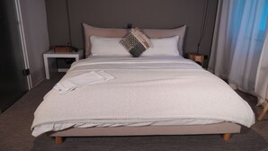 Un lit bien fait, recouvert d’une couverture blanche à texture intéressante, avec une serviette blanche pliée et un coussin décoratif.