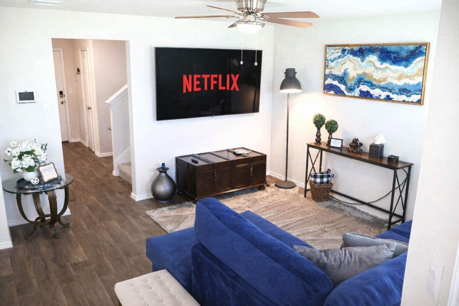 Una sala de estar con un sofá azul, un televisor montado en la pared, una lámpara de pie, un cuadro y un mueble aparador.